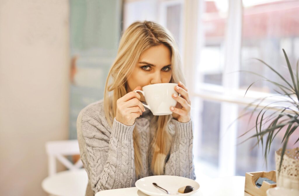 A woman sipping a hot beverage in a cozy indoor setting, wearing a gray sweater.