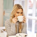 A woman sipping a hot beverage in a cozy indoor setting, wearing a gray sweater.