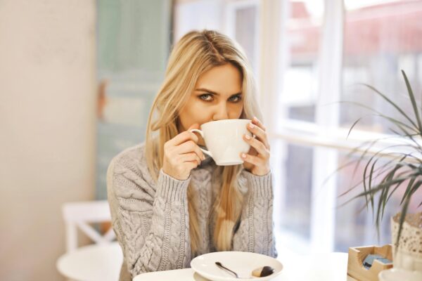 A woman sipping a hot beverage in a cozy indoor setting, wearing a gray sweater.